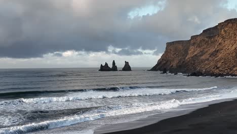 Scenic-footage-of-Vik's-black-sand-beach,-with-waves-crashing-and-the-iconic-Reynisdrangar-sea-stacks-in-the-distance