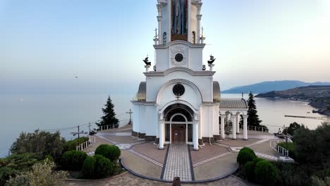 A-white-church-with-a-tall-spire-stands-on-a-hill-overlooking-the-sea-and-mountains,-as-the-sun-sets