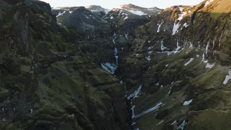 Aerial-landscape-view-over-a-mountain-canyon-with-a-river-flowing-and-a-frozen-waterfall