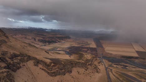 Aerial-view-of-the-landscape-at-Sólheimajökull,-capturing-the-dramatic-contrast-between-the-clear-and-stormy-skies,-with-rugged-terrain-and-a-distant-road-cutting-through-the-landscape