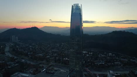Aerial-orbit-of-the-Costanera-tower,-mountains-with-the-sunset-in-its-final-stage,-Santiago-Chile