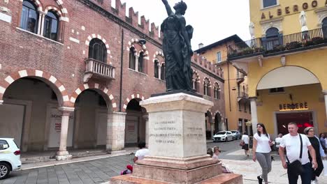 Square-with-liberty-sculpture-of-a-woman-with-people-walking-and-ancient-architecture,-red-brick-building-with-arches,-old-Verona-in-Italy