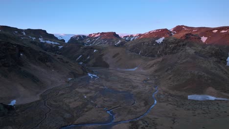 Aerial-landscape-view-of-melting-snow-river-flowing-down-mountains-and-glacial-valleys,-in-Iceland-at-dusk
