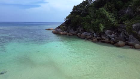 Turquoise-ocean-water-meeting-a-rocky-shore-with-a-lush-green-jungle-forest-on-a-tropical-island