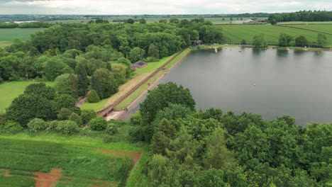 A-view-above-the-Kettering-Reservoir:-a-steady-descending-shot-capturing-vivid-bright-green-foliage-and-still-water-surface