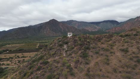 Aerial-view-of-the-top-of-the-mountain-in-Cafayate-with-a-religious-house-and-a-stunning-view-of-the-valley-and-farming-and-more-hills-at-the-horizon