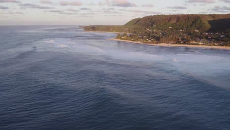 An-aerial-view-of-the-stunning-North-Shore-coastline-in-Hawaii,-showcasing-the-blend-of-ocean-waves,-sandy-beaches,-and-lush-green-hills-under-a-soft-evening-light