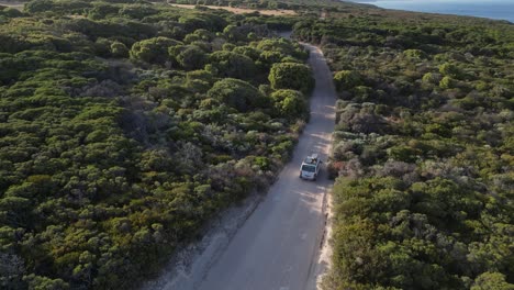 White-van-driving-on-dusty-road-at-sunset-surrounded-by-trees,-Western-Australia,-aerial