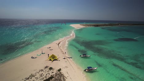 A-tropical-beach-with-kitesurfers-and-boats-in-turquoise-waters-on-a-sunny-day,-aerial-view