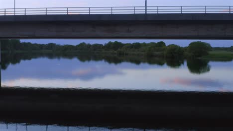 Aerial-dolly-below-bridge-spanning-River-Corrib-reflecting-soft-pastel-blue-golden-sky-and-vegetation