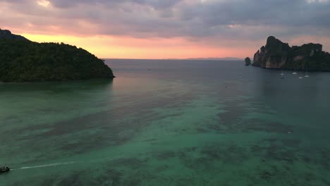 Schöner-Luftbildflug-über-Eine-Tropische-Insel-Bei-Sonnenuntergang-Und-Wolkigem-Himmel-Mit-Booten,-Die-Auf-Einem-Türkisfarbenen-Meer-Segeln