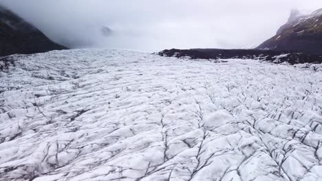 Vuelo-De-Proximidad-Ascendente-Sobre-Un-Glaciar-En-Islandia,-Destacando-Los-Impresionantes-Patrones-De-Hielo-Polar,-Las-Montañas-Y-Un-Cielo-Nublado-En-El-Horizonte.