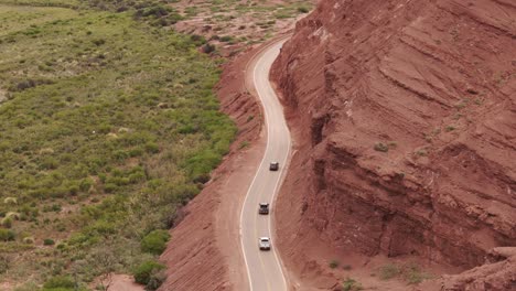 Vista-Aérea-De-La-Carretera-En-El-Valle-Calchaquí,-Con-Rocas-Rojas-A-La-Derecha-Y-Pasto-Verde-Silvestre-A-La-Izquierda,-Mientras-Los-Autos-Circulan-Por-El-Valle.