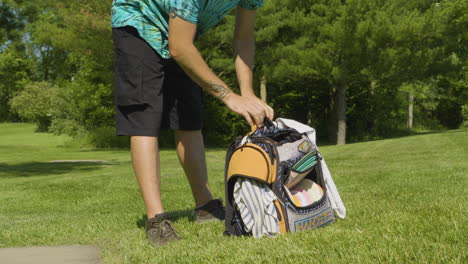 A-man-organizes-his-disc-golf-bag,-ensuring-everything-is-in-place-for-the-game,-set-in-a-sunny-park-with-lush-greenery