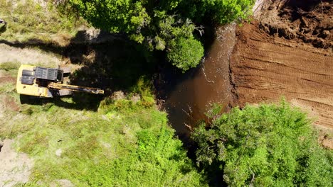 Aerial-Birds-Eye-shot-of-an-excavator-digging-near-a-riverbed-surrounded-by-lush-greenery-in-Chiloe,-Chile
