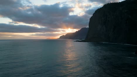 Drone-footage-flying-over-the-sea-in-Madeira-with-mountains-in-the-background-and-a-beautiful-sunrise-breaking-through-the-clouds