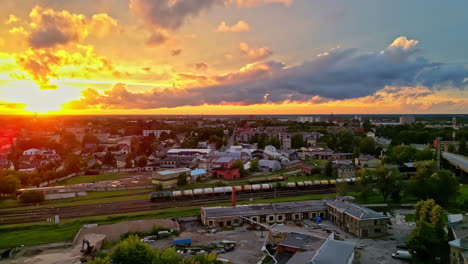 Drone-shot-in-slowmotion-sunset-over-a-city-with-rail-road-and-a-train-and-green-places-between-the-houses-in-Latvia,-Jelgava