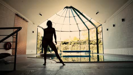 Silueta-De-Mujer-Acróbata-Haciendo-El-Pino-Y-La-Pierna-Posando-Junto-A-La-Piscina-Cubierta