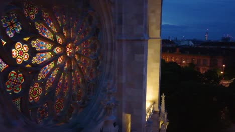 Vista-Nocturna-De-Un-Rosetón-De-Vidrieras-En-Una-Iglesia-Gótica-En-Viena,-Austria,-Con-Las-Luces-De-La-Ciudad-Al-Fondo