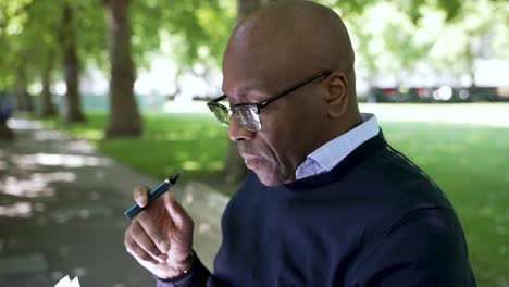 Bald-Black-male-vaping-while-sitting-outdoors-on-a-bench-in-a-park