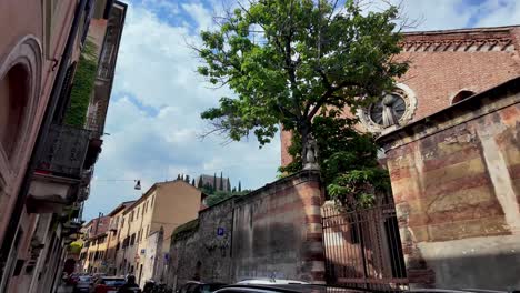 Hervorstehende-Obere-Teile-Der-Kirche-über-Hohe-Mauer-Mit-Einem-Baum-Und-Vorbeifahrenden-Autos-Auf-Der-Straße-In-Verona