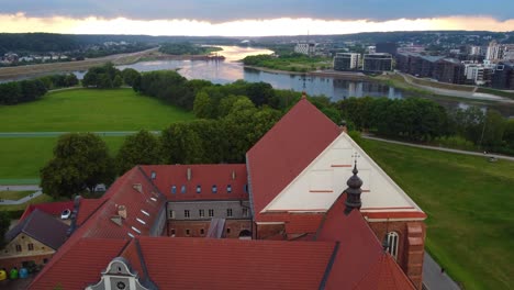 Vista-Aérea-De-Un-Edificio-En-Kaunas,-Lituania,-Con-Tejados-Rojos,-Campos-Verdes-Y-Un-Río-Al-Fondo.