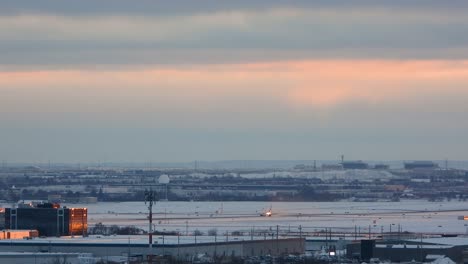 Winter-view-of-an-airport-in-Toronto,-Canada,-with-snow-covered-runways-and-a-plane-taking-off-or-landing-during-sunset