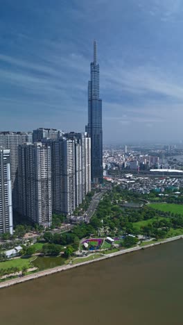Vertical-hyperlapse-of-Landmark-building-and-Central-park-along-the-Saigon-River-on-beautiful-sunny-day-with-blue-skies-showing-impressive-architecture-and-landscaping-with-container-boat-on-river