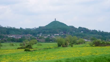 Scenic-view-of-The-Tor-in-rural-countryside-of-Glastonbury-overlooking-green-fields-of-wild-yellow-flowers-on-the-Somerset-Levels-in-England-UK