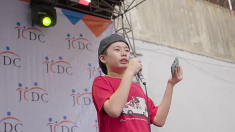 Young-boy-with-autism-sings-at-outdoor-JCDC-event-holding-microphone-and-phone,-Jakarta-Barat,-Indonesia