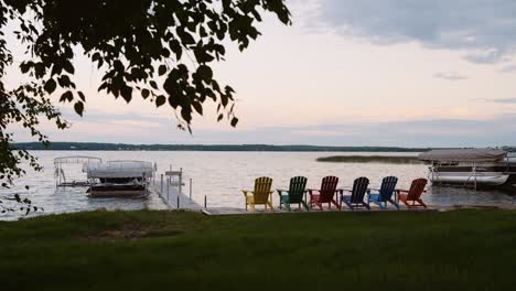 Sechs-Farbenfrohe-Adirondack-Stühle,-Ordentlich-Aufgestellt,-Mit-Blick-Auf-Einen-See-Bei-Sonnenuntergang-Mit-Einem-Dock-Und-Einem-Bootslift-Im-Wasser