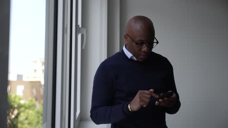 Bald-Black-man-stands-by-a-window-in-a-modern-room,-focused-on-his-smartphone