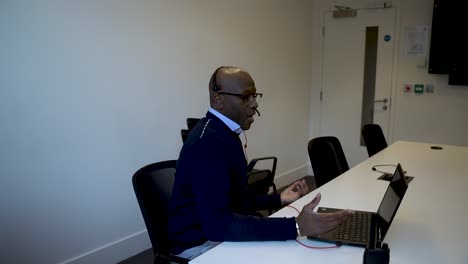 Black-male-professional-putting-on-headset-while-seated-in-a-meeting-room-with-a-laptop-and-taking-call