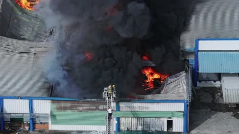 Vista-Aérea-De-Teleobjetivo-De-Los-Bomberos-En-La-Escalera-Extinguiendo-Llamas-Con-Manguera-En-El-Almacén-En-Llamas