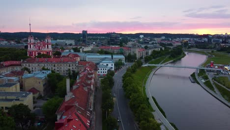 Aerial-view-of-Vilnius,-Lithuania,-at-sunset,-featuring-the-river,-bridges,-buildings,-and-a-prominent-red-church