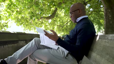 Bald-Black-Man-relaxing-on-a-bench-during-a-lunch-break,-vaping-and-reading-under-bright-green-trees-in-a-park