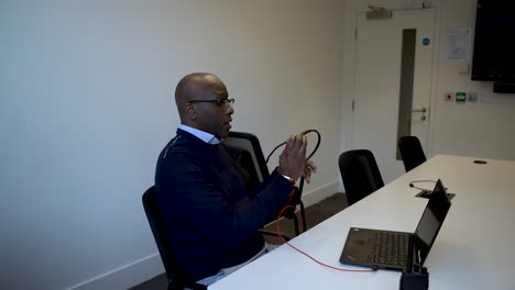 Un-Hombre-Negro-Desenredando-Los-Auriculares-Mientras-Está-Sentado-En-Una-Sala-De-Reuniones-Con-Una-Computadora-Portátil