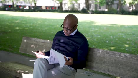 Un-Hombre-Está-Sentado-En-Un-Banco-De-Madera-En-Un-Parque,-Leyendo-Periódicos-En-Un-Día-Luminoso-Y-Soleado,-Con-Un-Exuberante-Césped-Verde-Al-Fondo,-Lo-Que-Sugiere-Serenidad-Y-Concentración