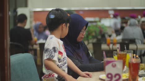 Una-Mujer-Indonesia-Con-Hijab-Alimenta-A-Un-Niño-En-Un-Restaurante-De-Comida-Rápida,-Disparada-A-Través-Del-Cristal-De-Una-Ventana,-Un-Centro-Comercial-Revela-La-Cultura-Moderna-Del-Sudeste-Asiático