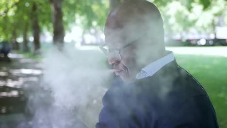 Bald-Black-Male-sitting-outdoors-in-a-park,-wearing-a-dark-sweater-and-glasses,-as-he-exhale-vapor-clouds-from-vaping