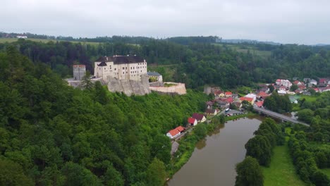 Aerial-view-of-Cesky-Sternberk-Castle-in-the-Czech-Republic,-surrounded-by-lush-greenery-and-a-river-with-a-small-village-nearby