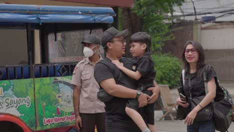 Indonesian-father-carrying-child-with-special-needs-near-colorful-odong-odong-bus-during-outdoor-event