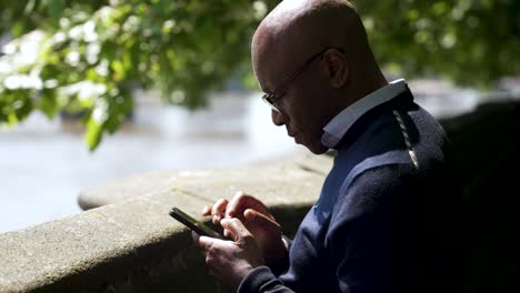 Black-male-standing-beside-a-wall-next-to-a-riverside,-using-a-smartphone