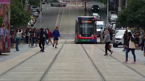 Melbourne-urban-lifestyle-captures-in-slow-motion-shot,-tram-running-along-Bourke-street,-passing-through-a-bustling-city-with-pedestrians-crossing,-commuters-waiting-at-the-stop