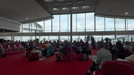 Patiently-sitting-people-waiting-for-plane-departure-by-reading-and-using-their-mobiles-at-Charles-de-Gaulle-Airport-Terminal-2-hall-in-Paris,-France