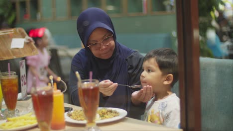 Portrait-Of-An-Indonesian-Mother-Wearing-A-Hijab-Spoon-Feeding-Her-Young-Son-In-A-Restaurant