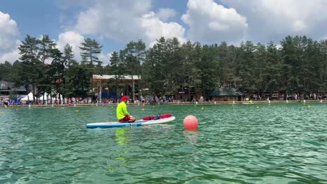 Young-man-in-a-neon-yellow-shirt-and-red-cap-sits-on-a-paddleboard-in-a-calm,-green-lake