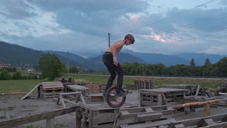 Young-male-unicyclist-balancing-on-wooden-piles-and-jumping-with-mountains-in-background-under-an-overcast-sky