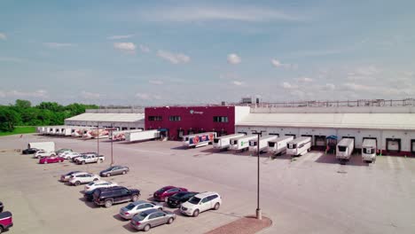 Vista-Aérea-De-Un-Linaje-De-Centro-De-Distribución,-Con-Camiones-Estacionados,-Muelles-De-Carga-Y-Un-área-De-Estacionamiento-Bajo-Un-Cielo-Despejado.