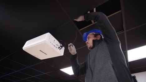 Worker-in-a-hardhat-installing-or-repairing-a-ceiling-panel-next-to-a-ceiling-mounted-projector